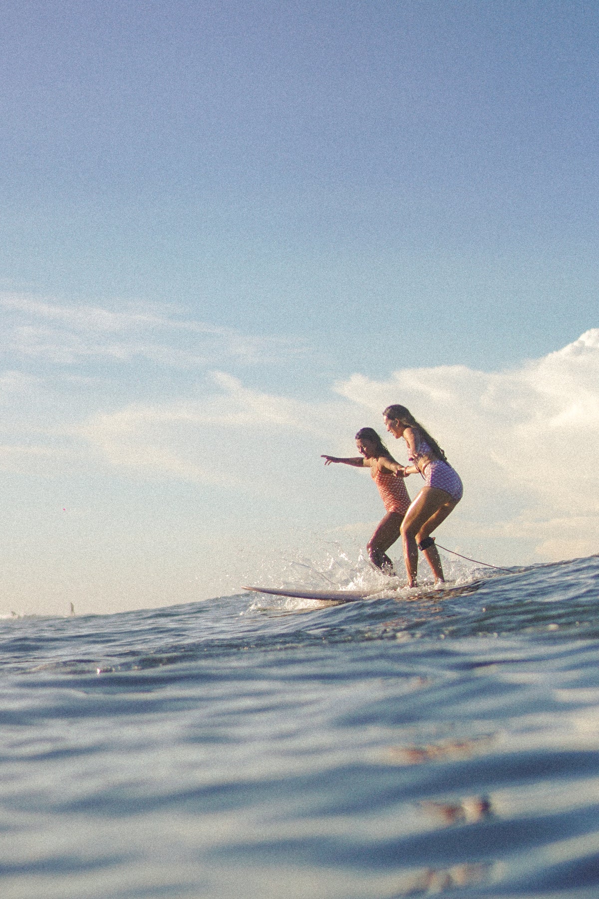 Two women surfing on a small wave under a clear blue sky, with the ocean water in the foreground and a soft vintage filter.slider_item_pzWWEb