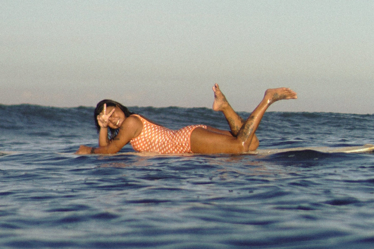 A woman in a orange and white patterned swimsuit lies on a surfboard on the ocean water under a clear blue sky.