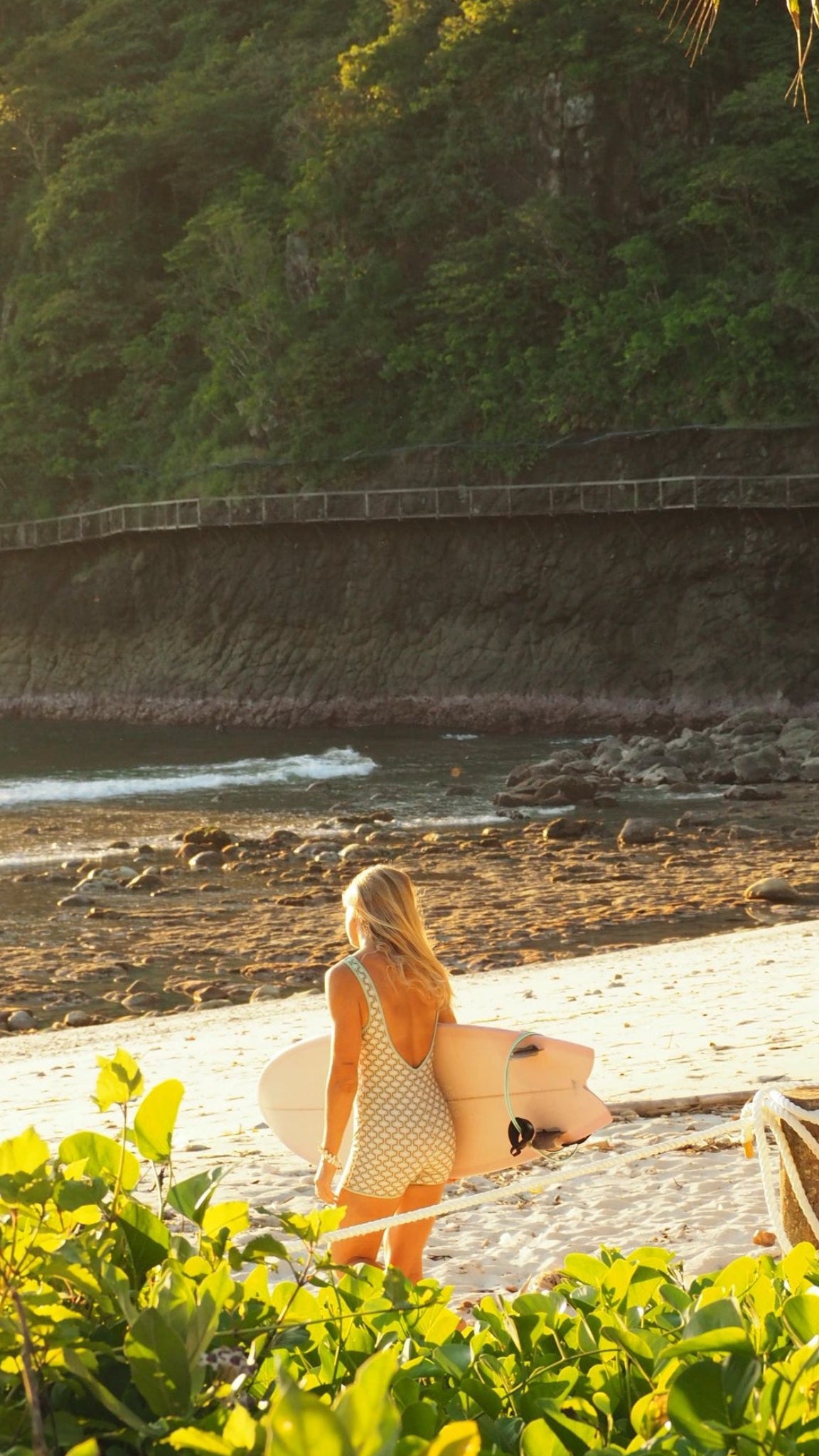 A woman in a beige dress carrying a surfboard walks onto a sandy beach with a rocky shoreline and lush green cliffs in the background, framed by bright green foliage in the foreground.